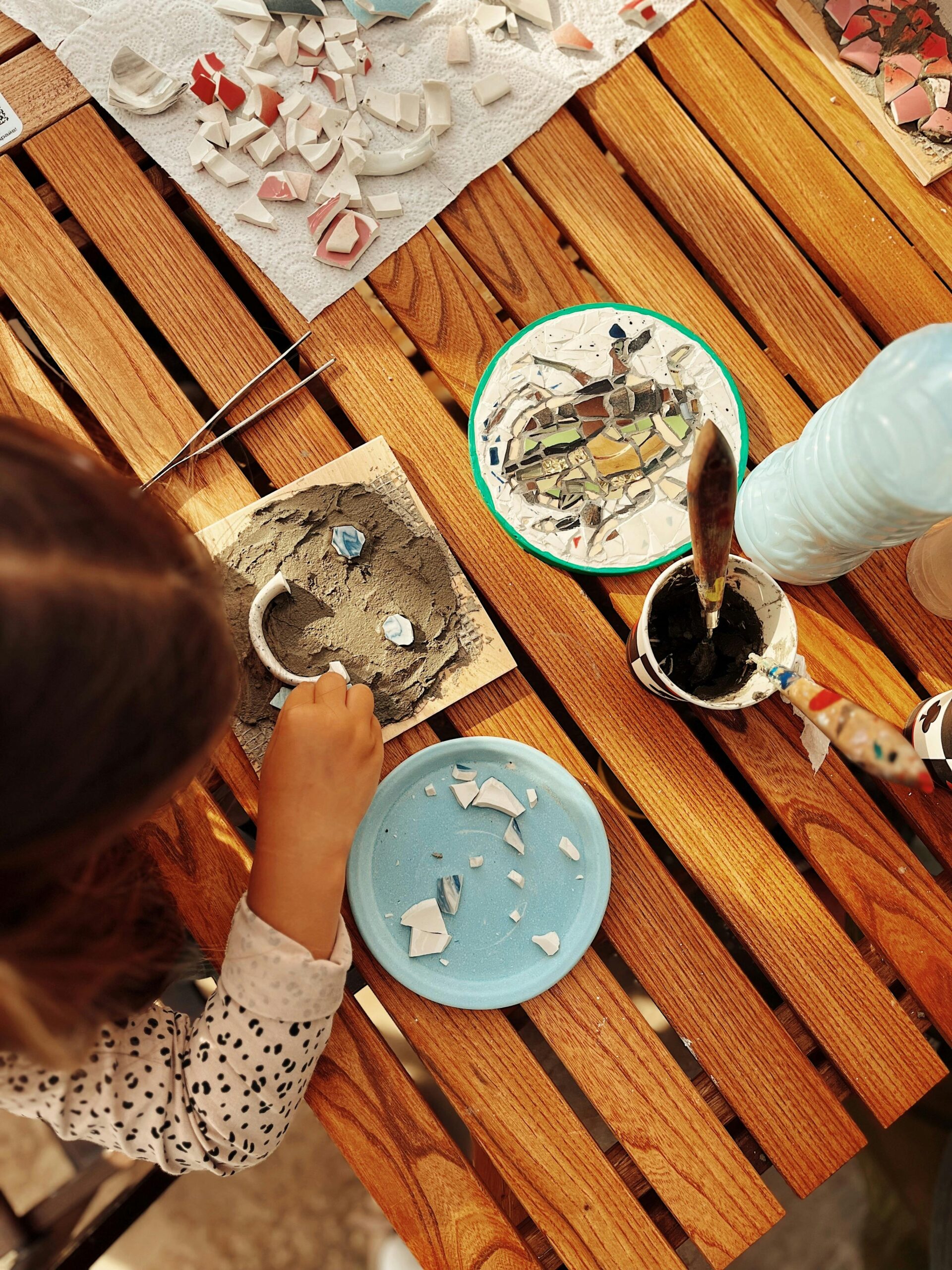 Top view of a child creating a mosaic art piece with colorful tiles on a wooden table.