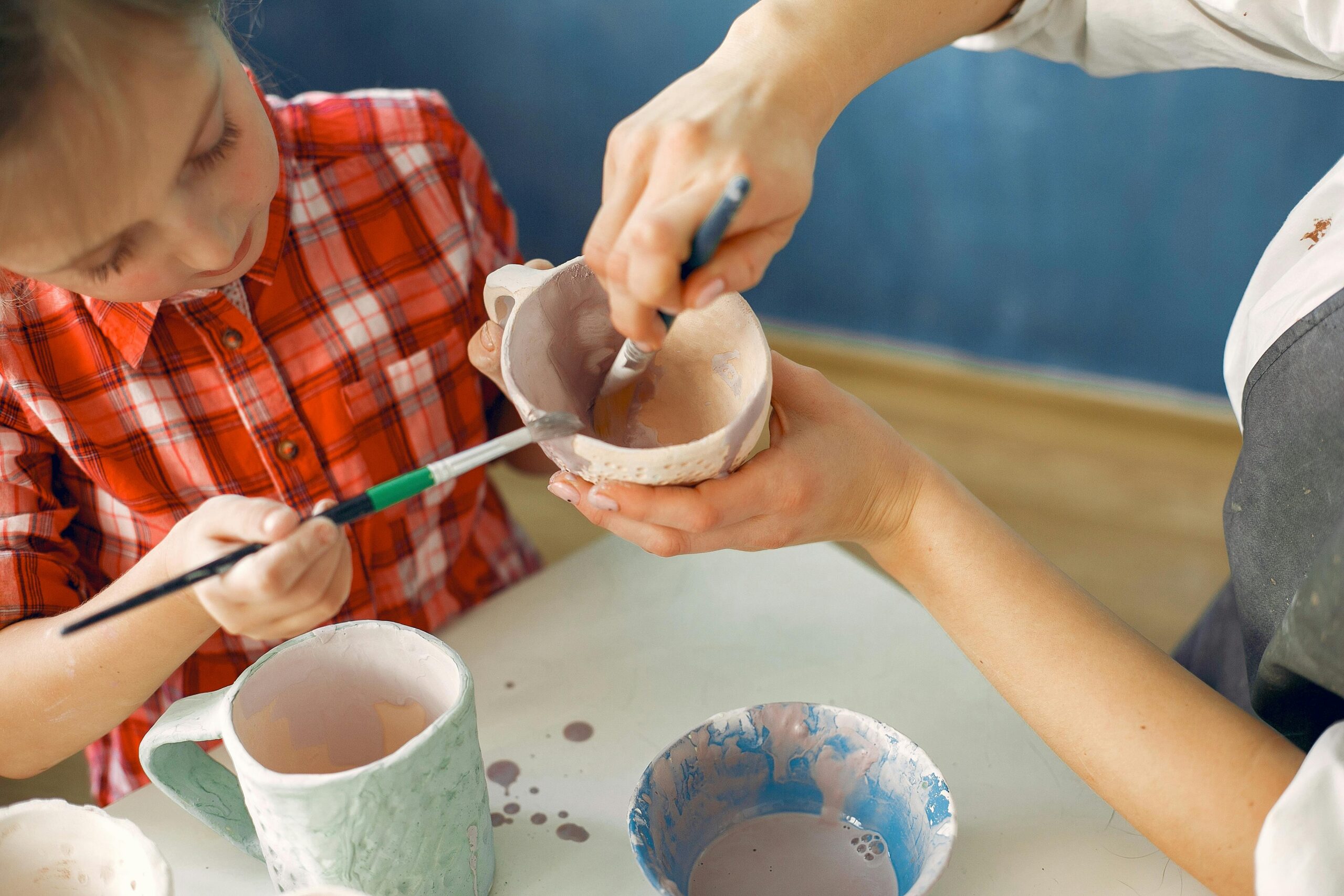 From above crop female ceramist and cute little girl painting handmade ceramic dishware while sitting at table together in creative pottery